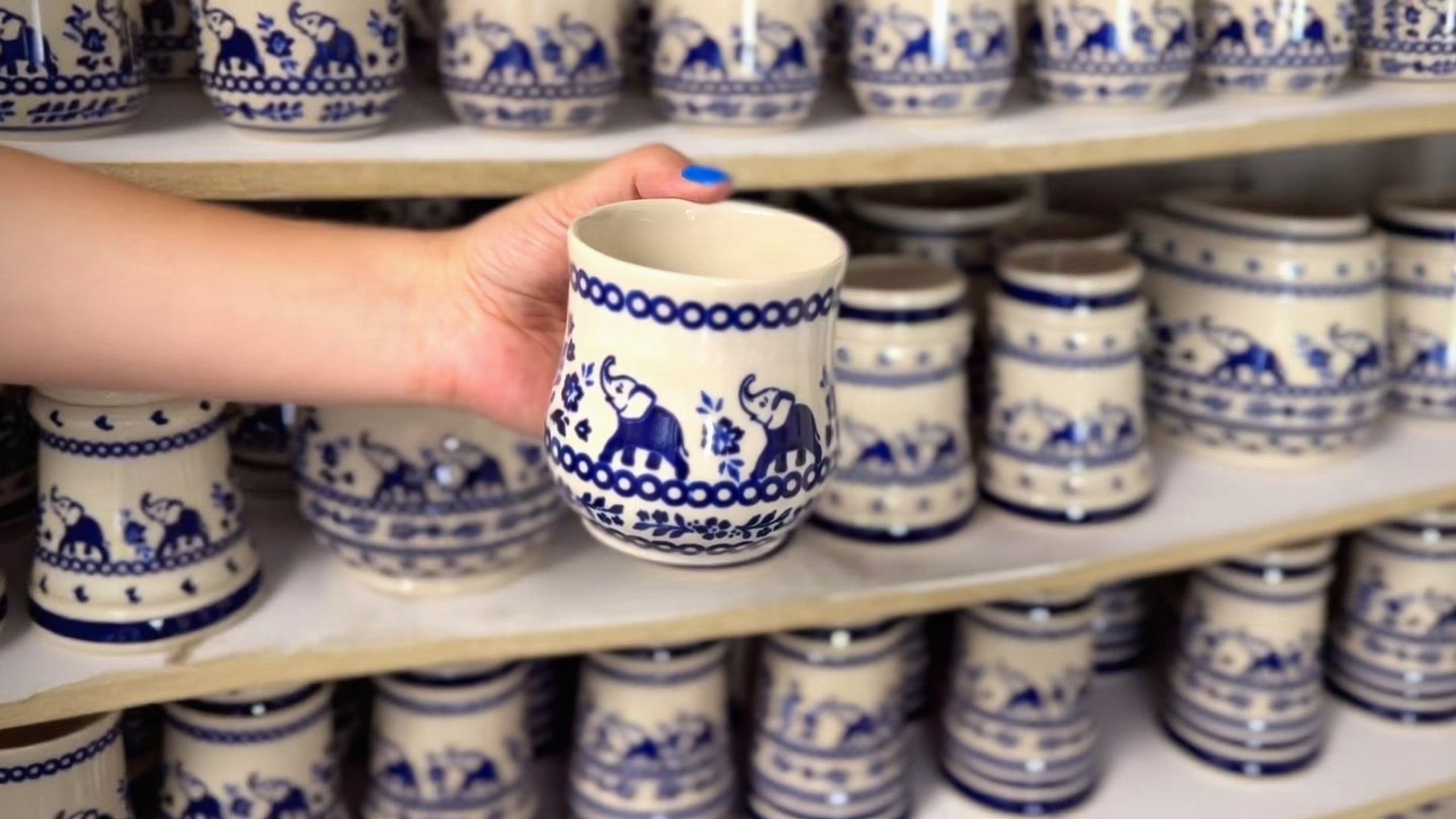 A Bolesławiec Elephants mug just out of the kiln, held in a hand, with shelves of mugs in the same pattern behind it